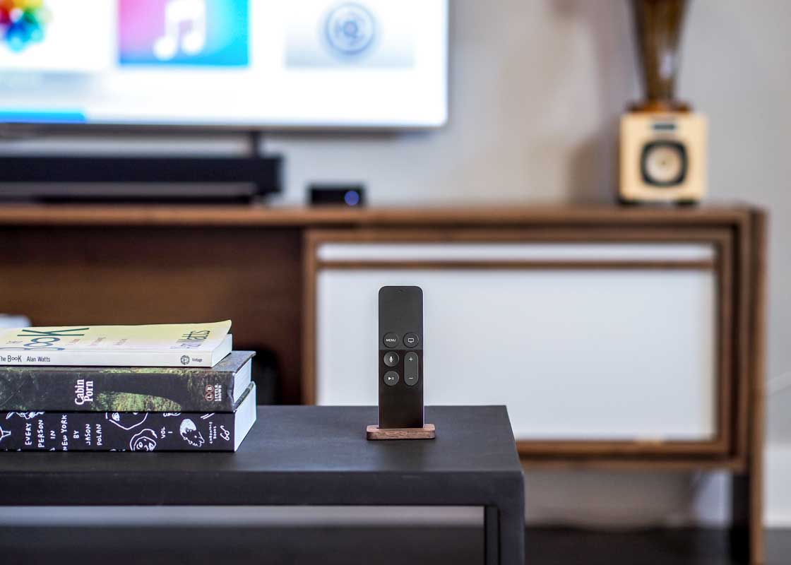 an Apple TV remote sitting in the stand on a coffee table infront of a tv.