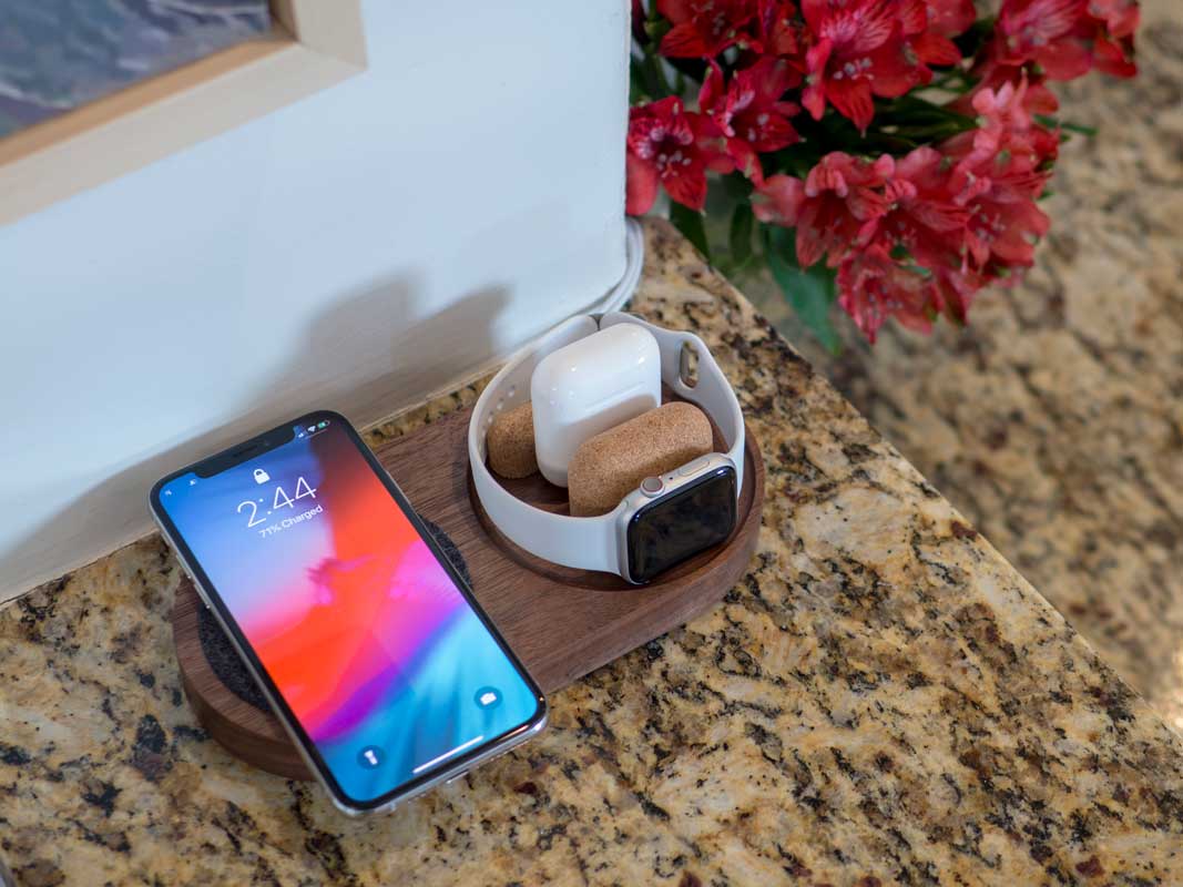 a material dock with a phone charging via qi, an Airpods case charging via cable, and an Apple Watch charing on it's side.